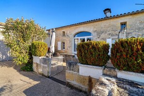 Un bâtiment en pierre avec un toit recouvert de tuiles, une entrée fermée par un portail et un ciel d’un bleu limpide.