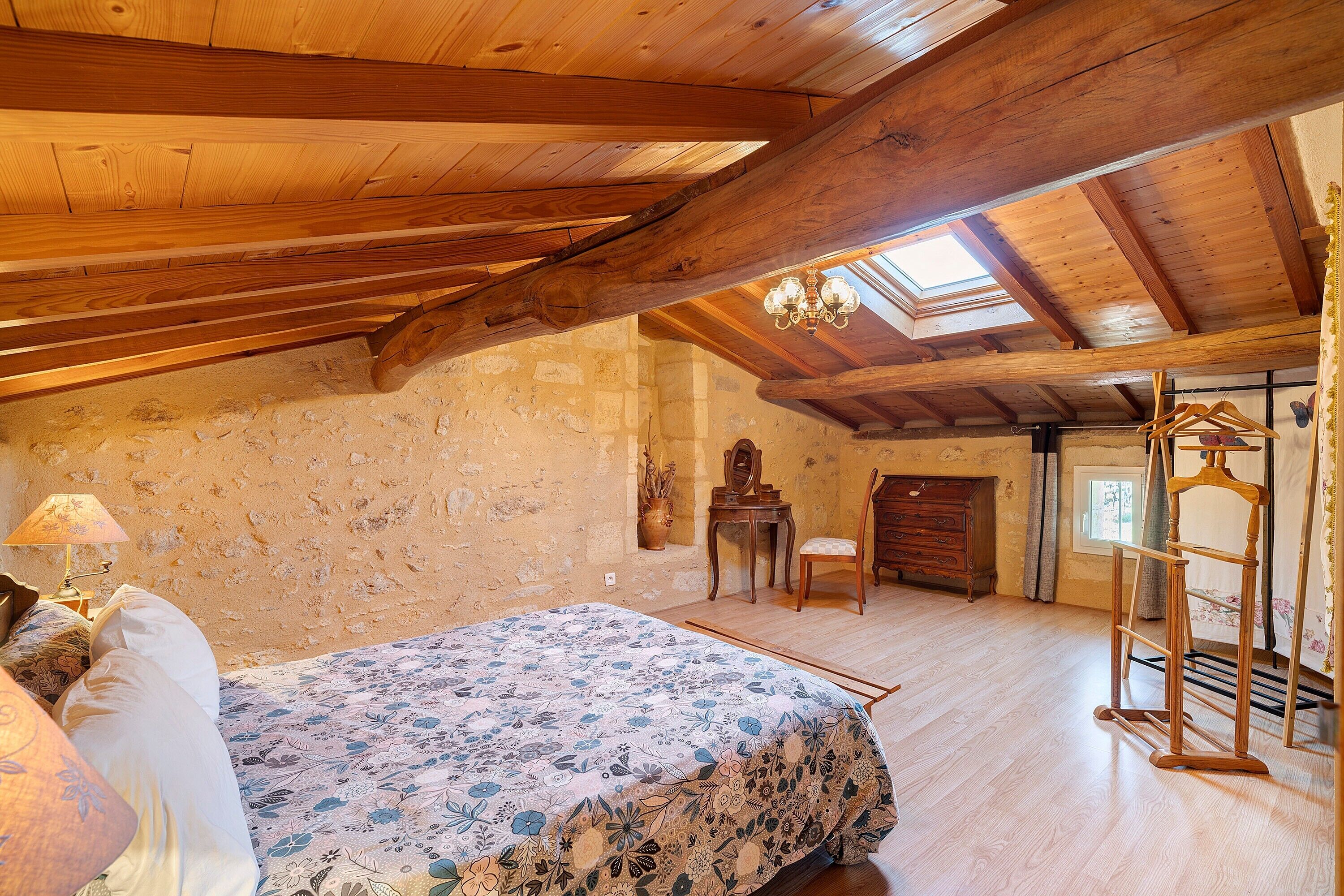 Une chambre avec un plafond en bois, un lit recouvert d’une couvre-lit à motifs floraux, une commode en bois et un puits de lumière.