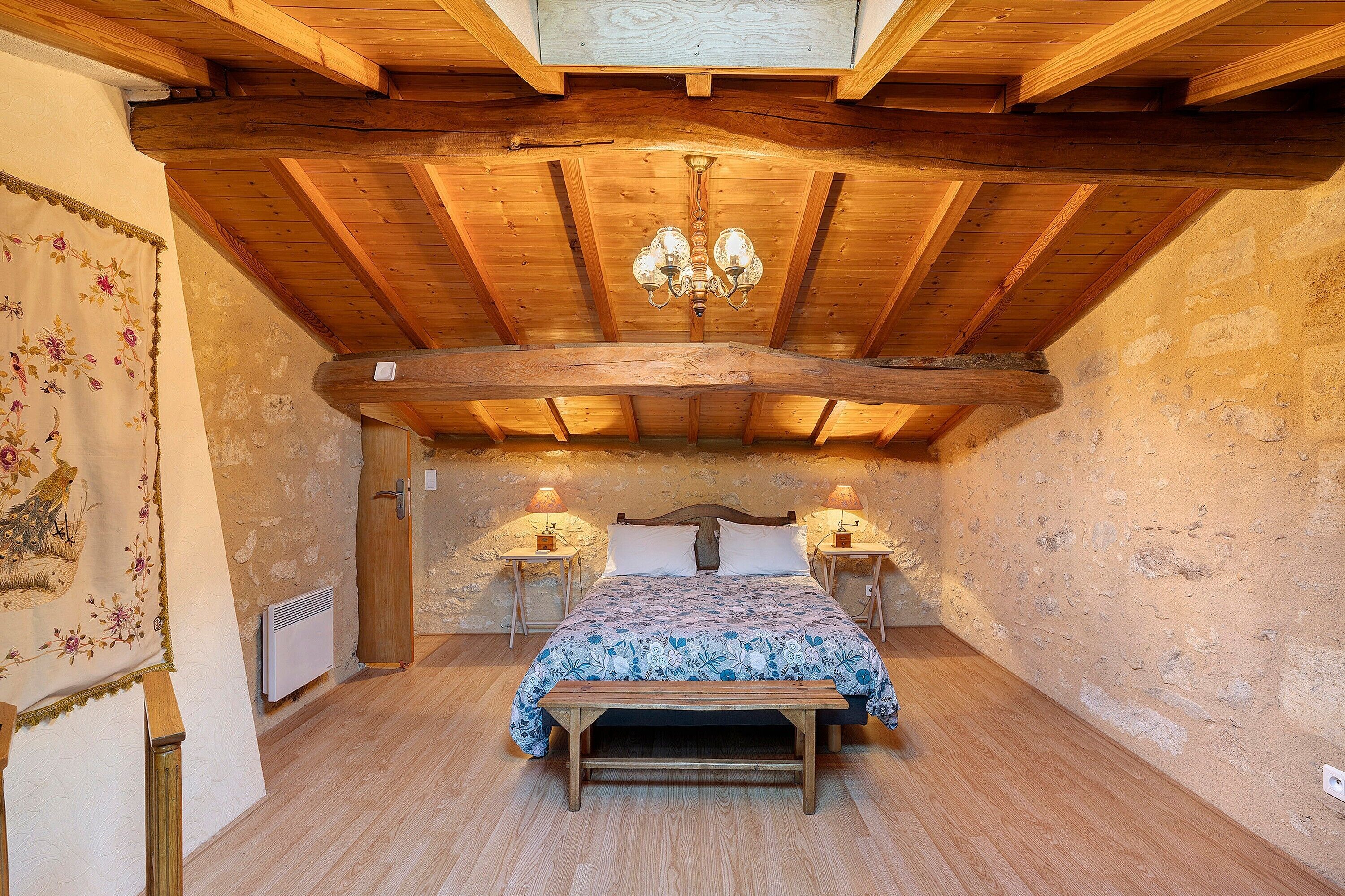 A bedroom with a wooden ceiling, a bed with a floral bedspread, two bedside lamps, and a wooden bench.