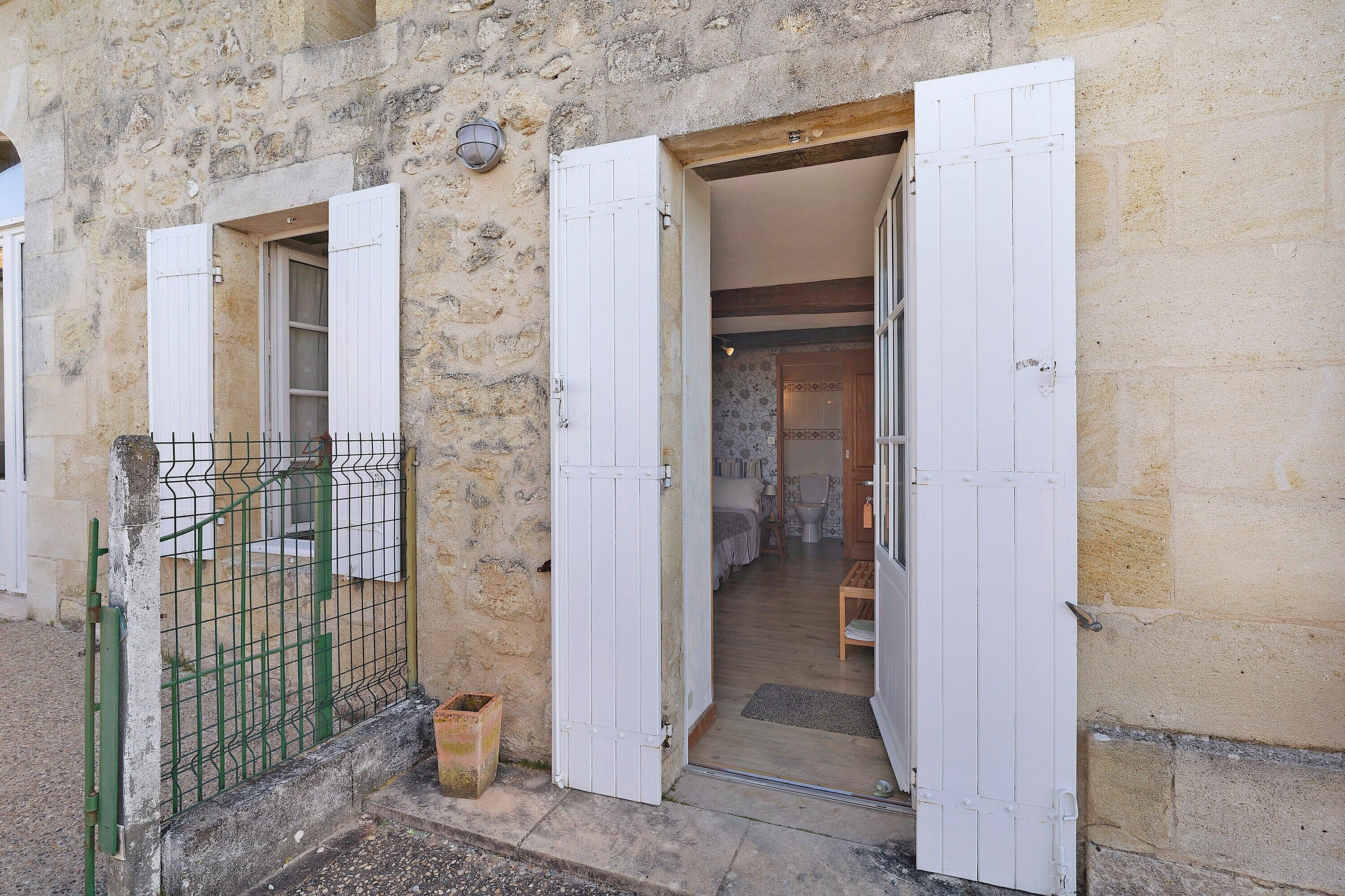 Un edificio de piedra con puerta y contraventanas de madera, una reja metálica y un interior visible con una cama y una silla.