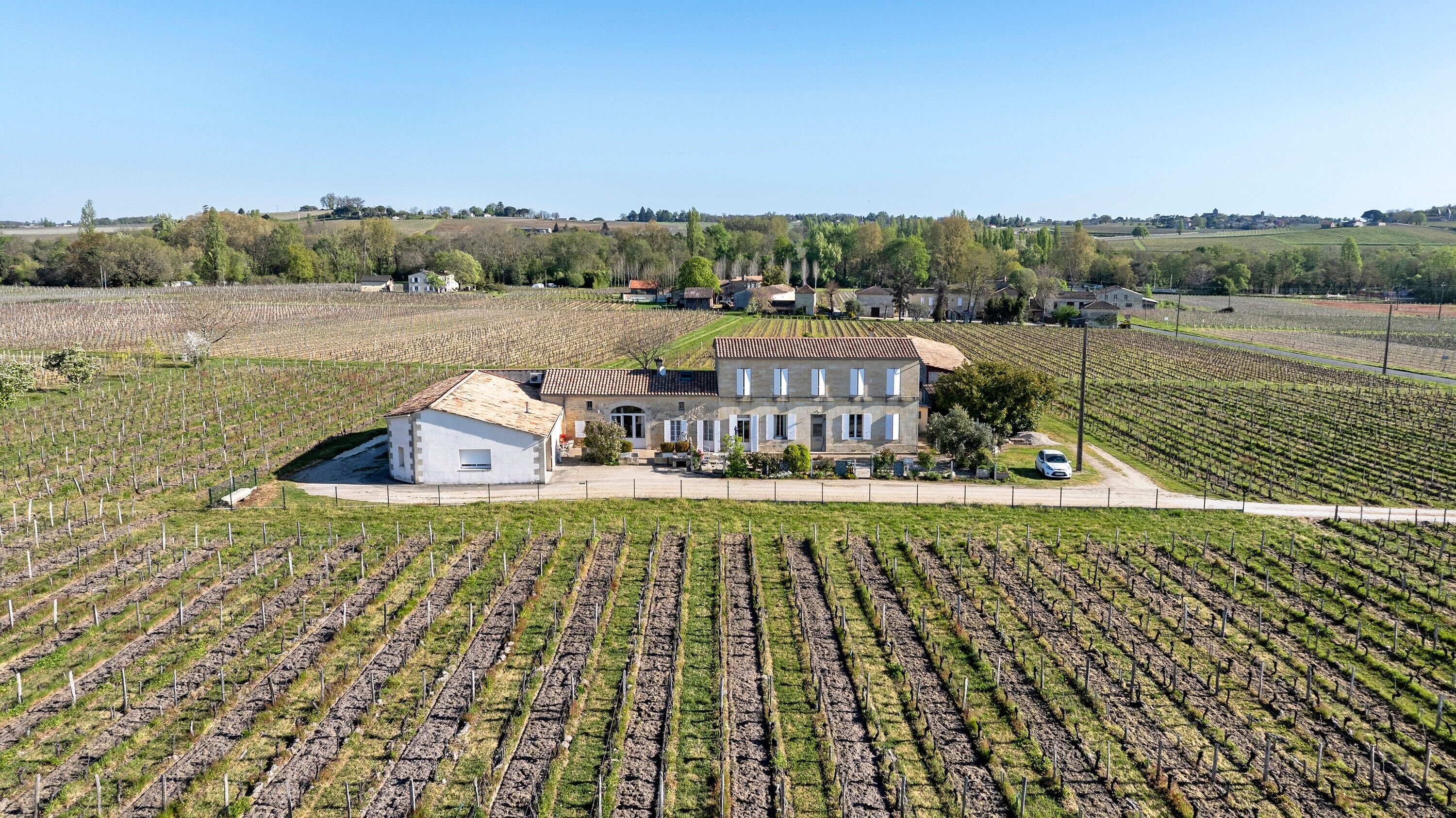 Un grand bâtiment à toit de tuiles, entouré de rangées de vignes, avec une construction plus petite située à sa gauche.