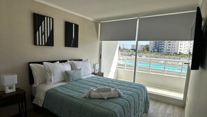 Une chambre avec un grand lit, une table de chevet et une fenêtre donnant sur une piscine et des immeubles.