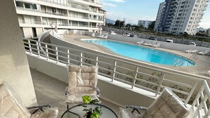 Un balcon doté d’une table en verre et de quatre fauteuils rembourrés, offrant une vue sur une piscine et des immeubles de grande hauteur.
