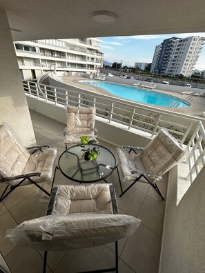 A balcony with a glass table and four cushioned chairs, overlooking a swimming pool and high-rise buildings. - Fabulous Apartment 2D2B Steps from the Beach - La Serena (La Serena)