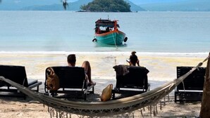 Uma praia com uma rede, duas cadeiras de descanso e um barco na água.