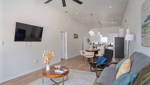 A modern living room with a flat-screen TV, a wooden coffee table, a sofa with cushions, a dining area with chairs, and a kitchen in the background. - Bright and Fancy 3-Bedroom Near Fun Riverwalk! (San Antonio)