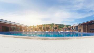 Ein Schwimmbecken mit Liegestühlen und Sonnenschirmen, umgeben von Gebäuden mit freiem Blick auf einen bewaldeten Hügel im Hintergrund.