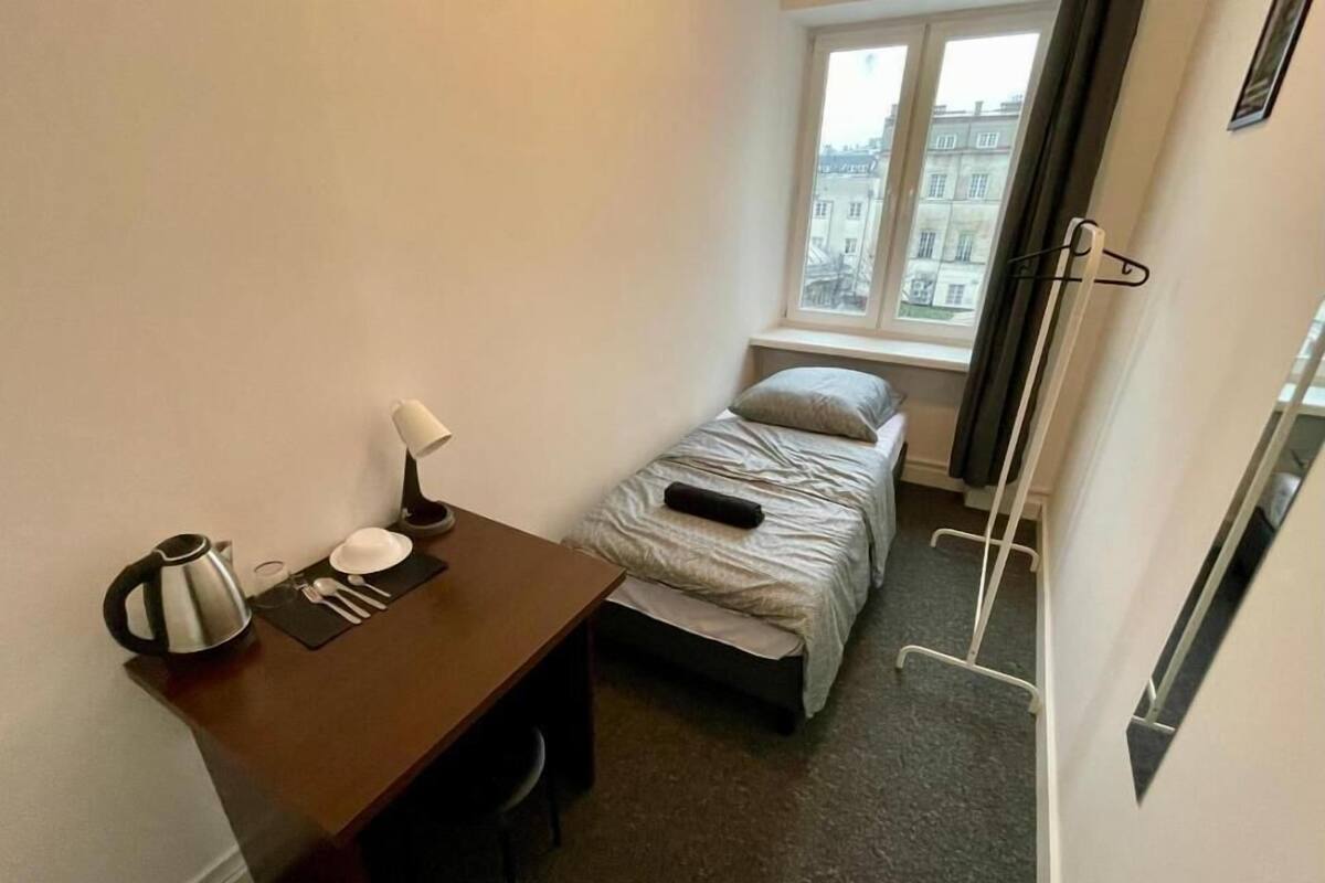 A small, compact room with a single bed, a desk with a kettle, a lamp, and a chair. There is a window providing a view of buildings outside.