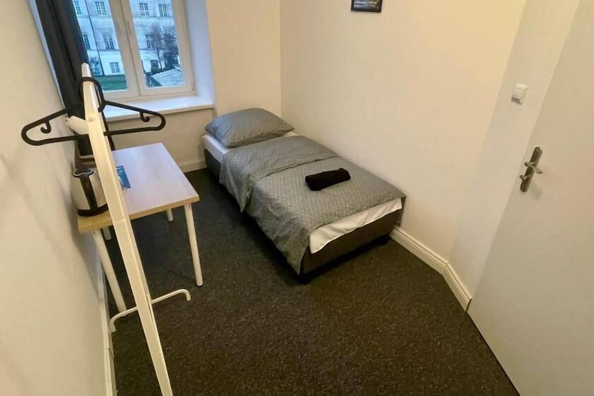 A small, compact room with a single bed, a desk, and a chair. There is a window providing a view of buildings outside.