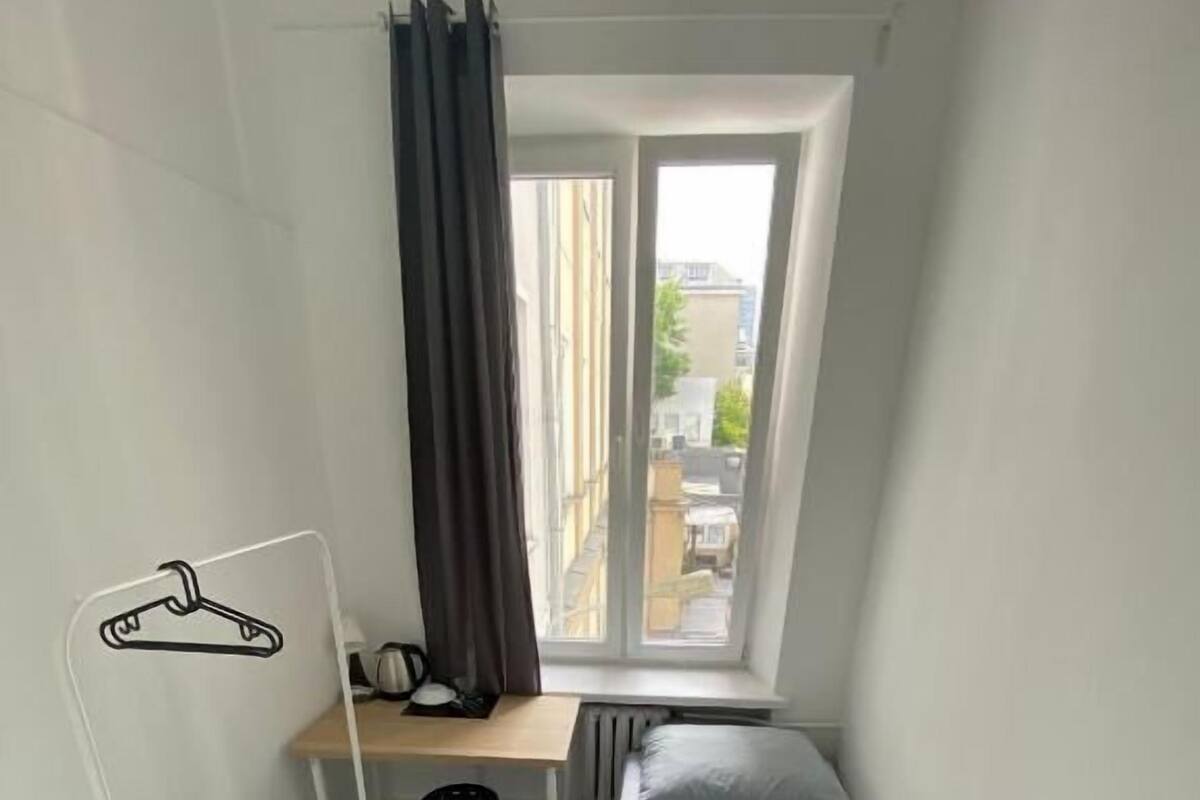 A small, well-lit room with a single bed, a desk, and a chair. There is a window with curtains, and a coat rack is visible.
