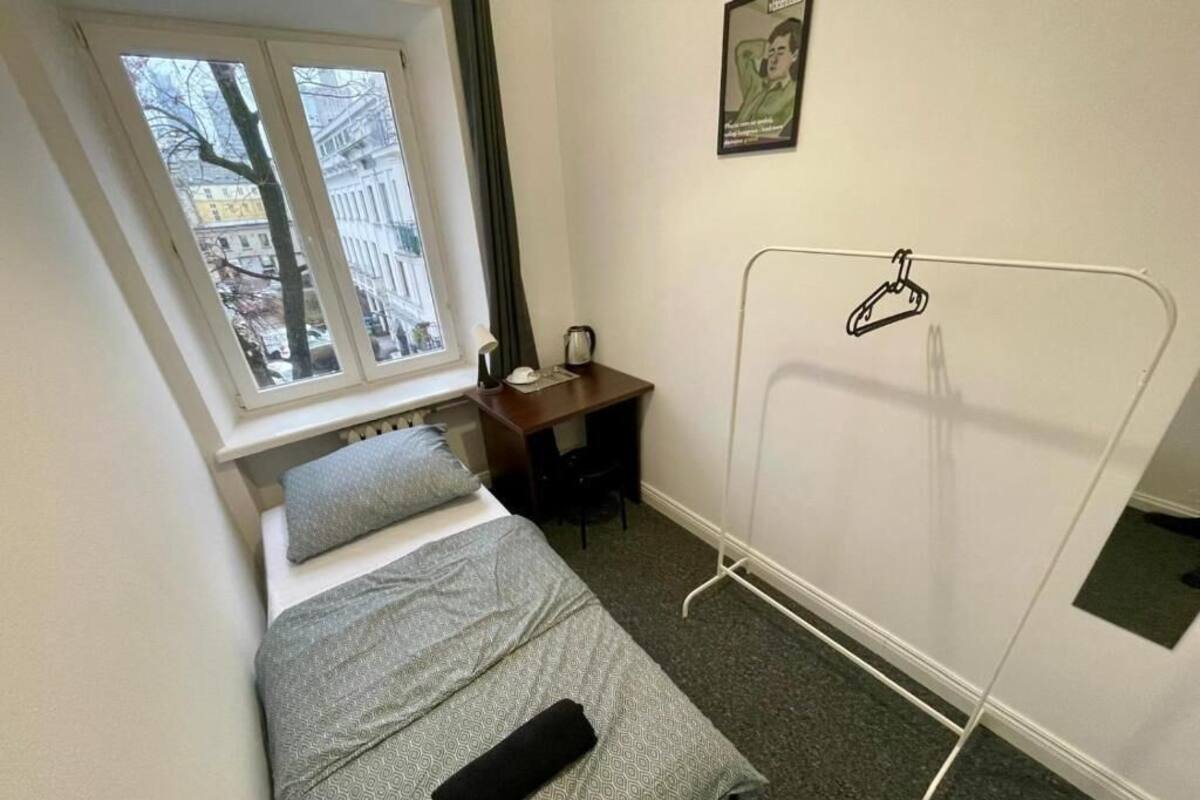 A small, neatly arranged bedroom with a bed, a desk, and a window providing a view of buildings outside.