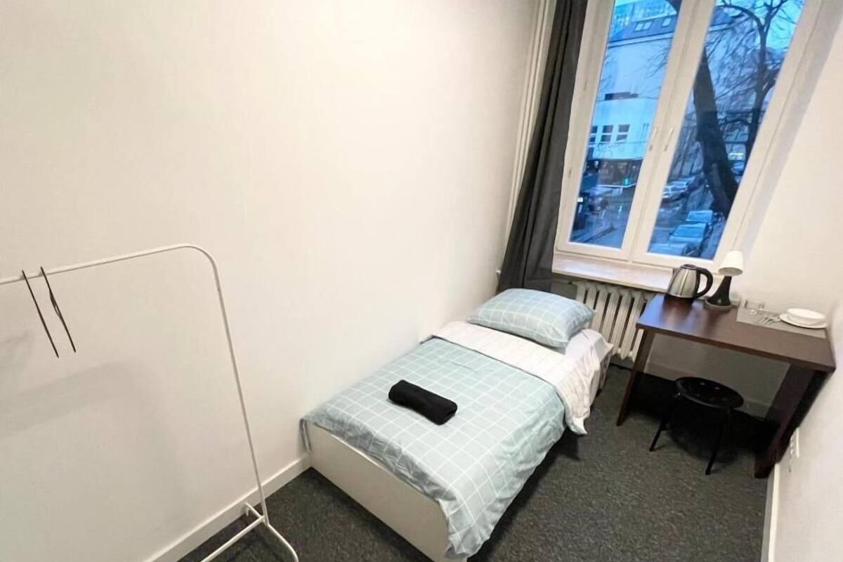 A small, neatly arranged bedroom with a bed, a radiator, a desk with a lamp, and a window with a view of buildings.