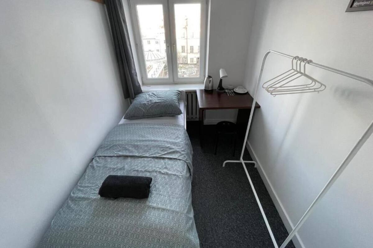 A small, compact room with a single bed, a desk with a chair, a radiator, and a window with curtains.