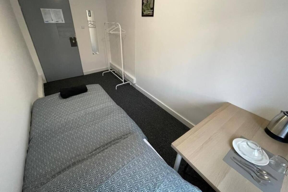 A small, single-bed room with a grey patterned bedspread, a wooden table with a kettle, and a framed picture on the wall.