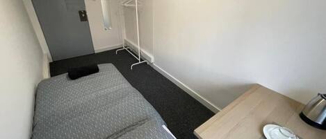 Une petite chambre avec un lit simple, une couverture à motifs gris, une table en bois avec une bouilloire et un tableau encadré accroché au mur.