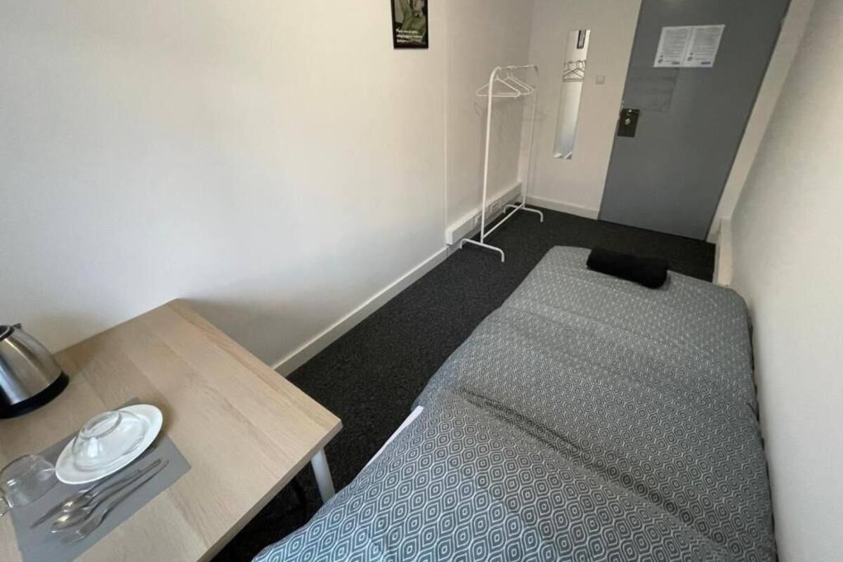 A small, compact hotel room with a single bed, a bedside table with a kettle, a small table with a plate and cutlery, a coat rack, and a door with a notice.
