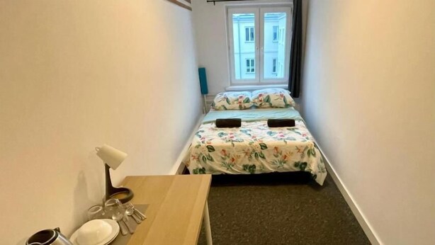 Una habitación pequeña con una cama al fondo, una mesita con una tetera, platos y vasos, y una ventana con cortinas.