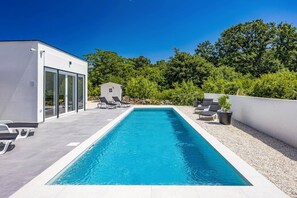 Una piscina rectangular con agua azul cristalina, rodeada por una terraza blanca y sillas de descanso negras.