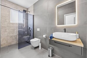 Un baño moderno con cabina de ducha de vidrio, inodoro blanco, lavamanos grande de color blanco con encimera de madera e iluminación integrada en el espejo.