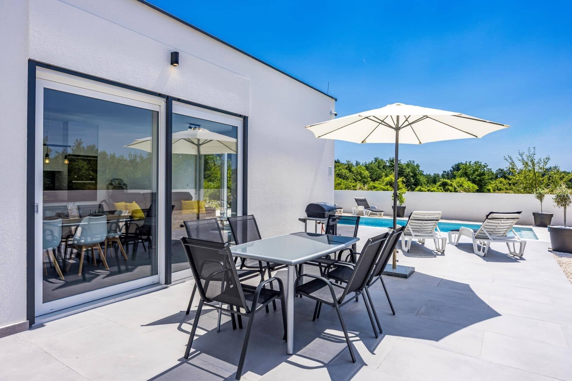 Un patio exterior con piscina, puertas de cristal y una zona de comedor con sillas y una mesa.
