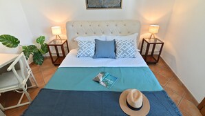 Une chambre à coucher bien rangée, avec un lit, des oreillers, un magazine et un chapeau posés sur le lit. On y trouve deux tables de chevet avec des lampes, et une grande plante placée à côté du lit.