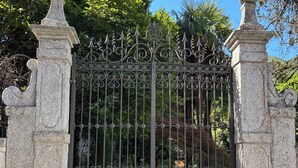 A wrought iron gate with intricate designs, flanked by stone pillars with rounded tops, set against a backdrop of lush greenery and a building with blue shutters. - The Lemon House Lemon House at the Lake. Elegant apartment with garden (Bellagio)