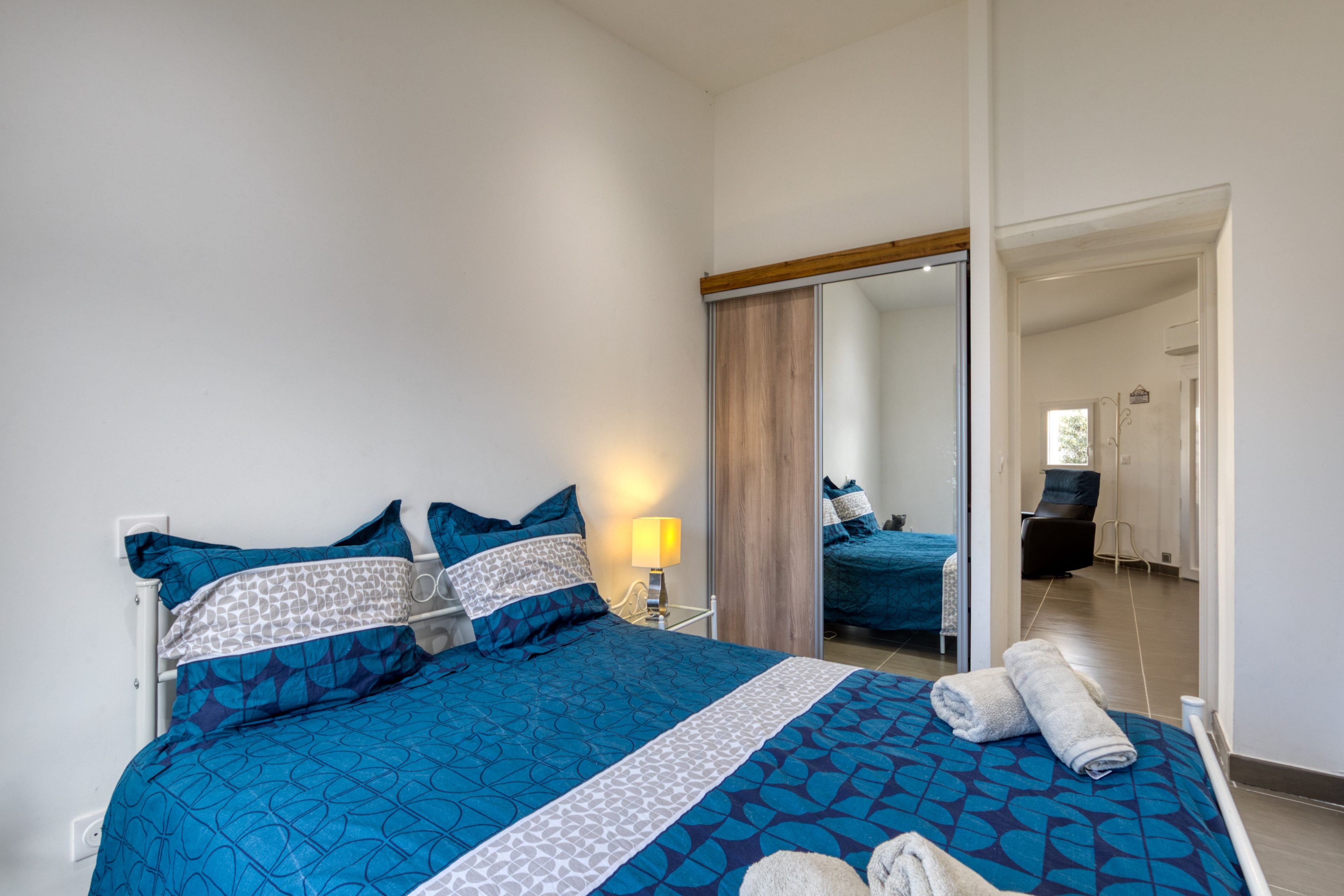 Un dormitorio con cama, almohadas, una mesita de noche, lámpara y espejo.