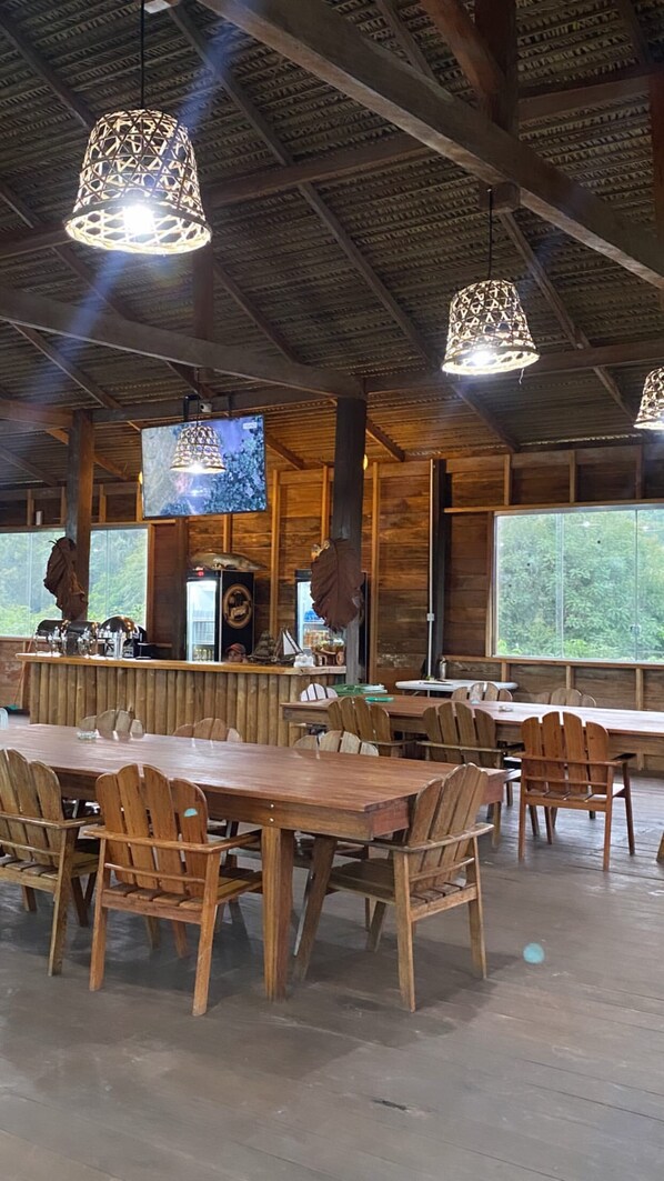 A spacious indoor dining area with wooden tables and chairs, hanging pendant lights, and large windows offering a view of the outdoors.