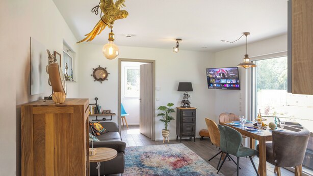 Een moderne woonkamer met een eethoek, een aan de muur bevestigde televisie en een bijzondere plafondlamp in de vorm van een vogel.