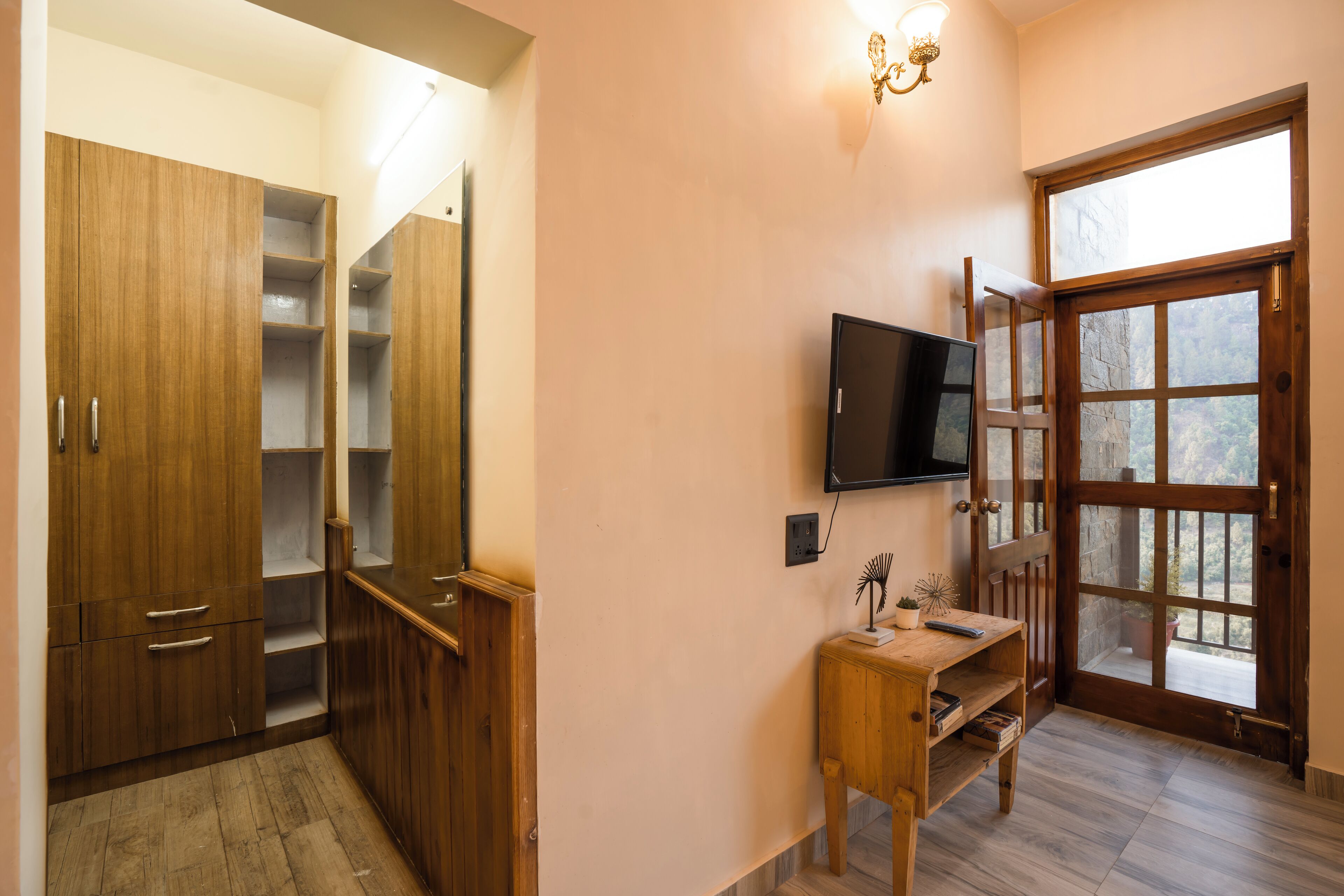 Una habitación con armarios de madera, un televisor montado en la pared, una pequeña mesa de madera y un ventanal con vistas a árboles.