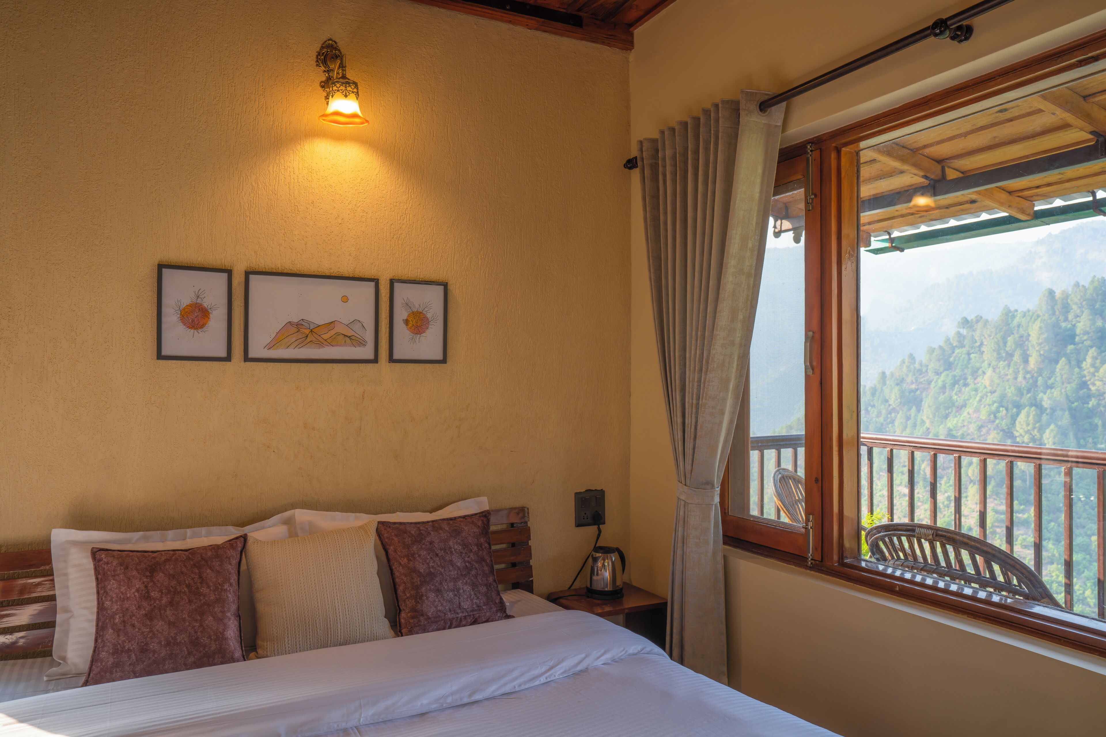 A bedroom with a bed, a nightstand with a lamp, a window with a view of a forest, and framed pictures on the wall.