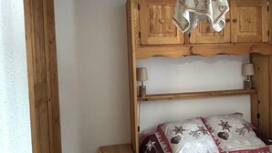 Ein Schlafzimmer mit einem hölzernen Kopfteil, einem Einzelbett mit weiß-roter Bettdecke, einem kleinen Holzschemel und einer Wandleuchte.