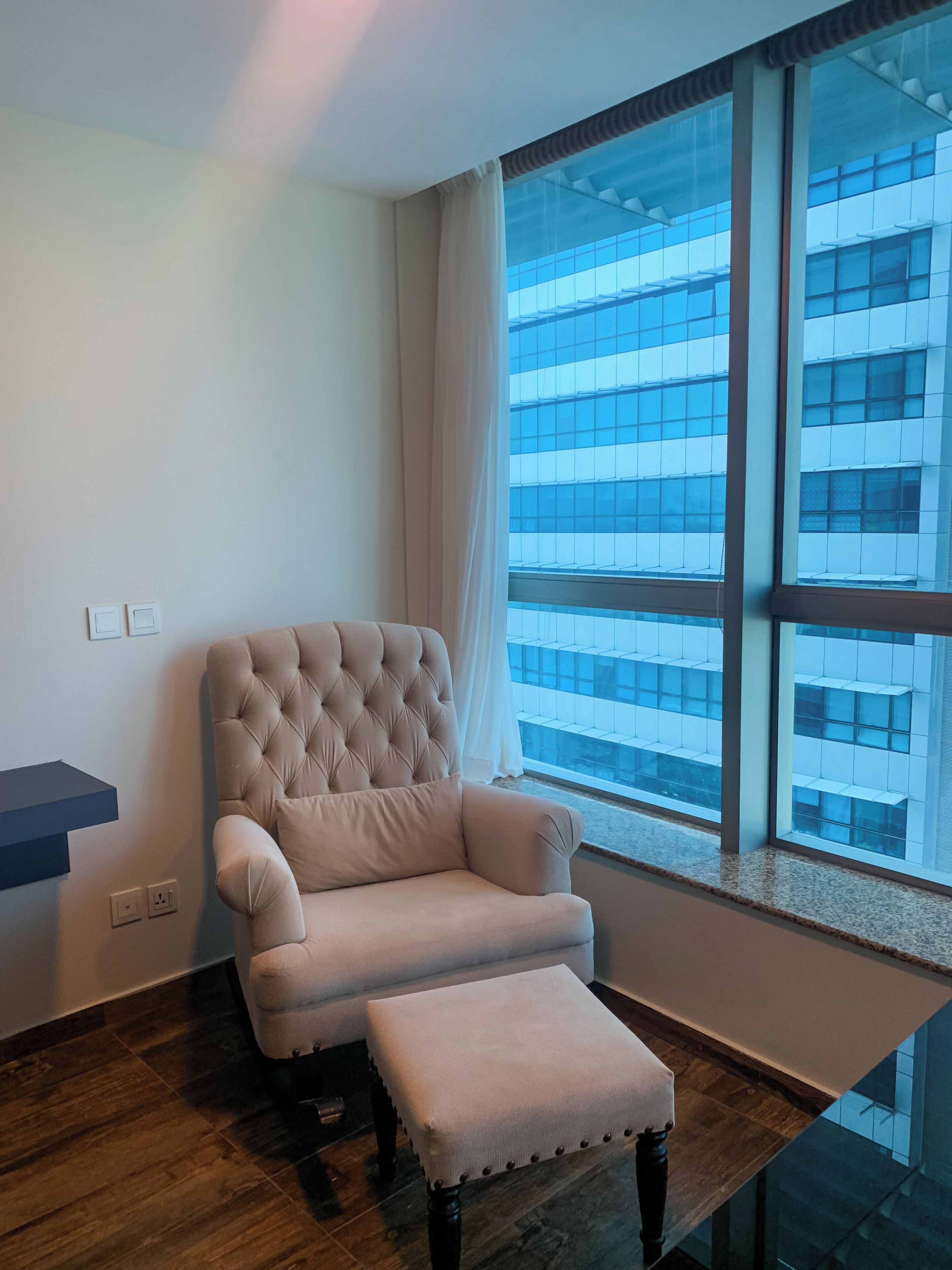 A modern room with a large window, a tufted armchair, a matching ottoman, and a dark wooden floor.