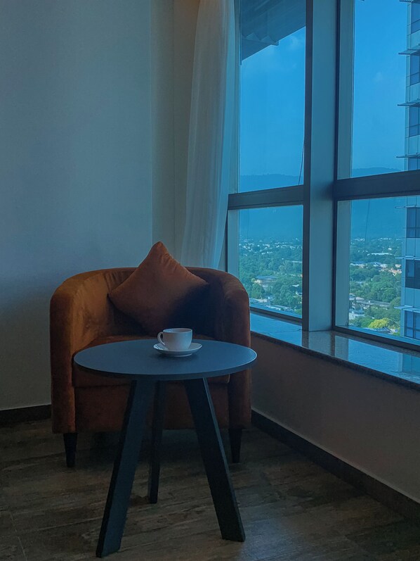 A room with a brown armchair, a small round table with a cup, and a large window offering a city view.