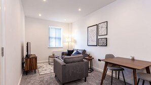A modern living room with a grey sofa, a wooden coffee table, and a dining table with chairs. There are abstract wall art pieces and a lamp on a side table. - Chic 1BR Bolton Apt near Manchester Rail (Bolton)
