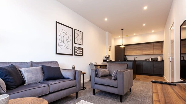 A modern living room with a grey sofa and armchair, a wooden coffee table, and a marble-patterned rug. A kitchen with dark wood cabinets and a microwave is visible in the background. - Chic 1BR Bolton Apt near Manchester Rail (Bolton)