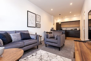 A modern living room with a grey sofa and armchair, a wooden coffee table, and a marble-patterned rug. A kitchen with dark wood cabinets and a microwave is visible in the background.