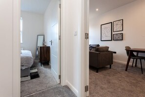 A bedroom with a bed, a dresser, and a mirror. There is a hallway leading to a living area with a couch and a table. - Chic 1BR Bolton Apt near Manchester Rail (Bolton)