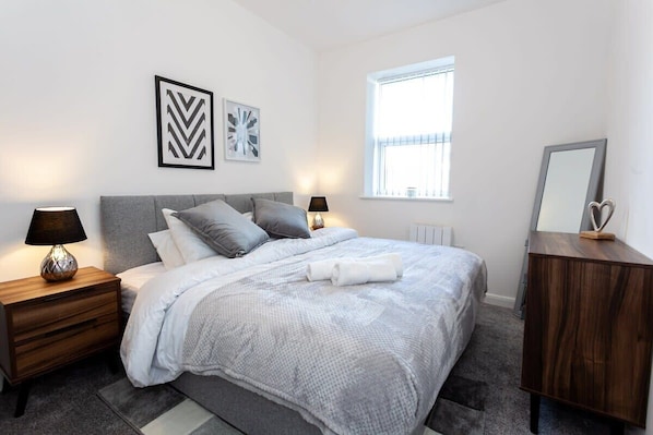 A modern bedroom with a large bed, two bedside tables with lamps, a dresser, and a window with blinds.