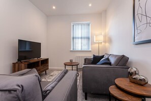 A modern living room with a flat-screen TV, grey sofas, a wooden entertainment unit, a small round table, and a floor lamp.