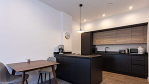 A modern kitchen with dark cabinetry, a central island, and a dining area with a table and chairs. - Chic 1BR Bolton Apt near Manchester Rail (Bolton)