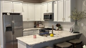 Una cocina moderna con armarios blancos, un refrigerador de acero inoxidable, un microondas y un lavamanos. Hay dos taburetes de bar y una silla de mimbre alrededor de la isla.