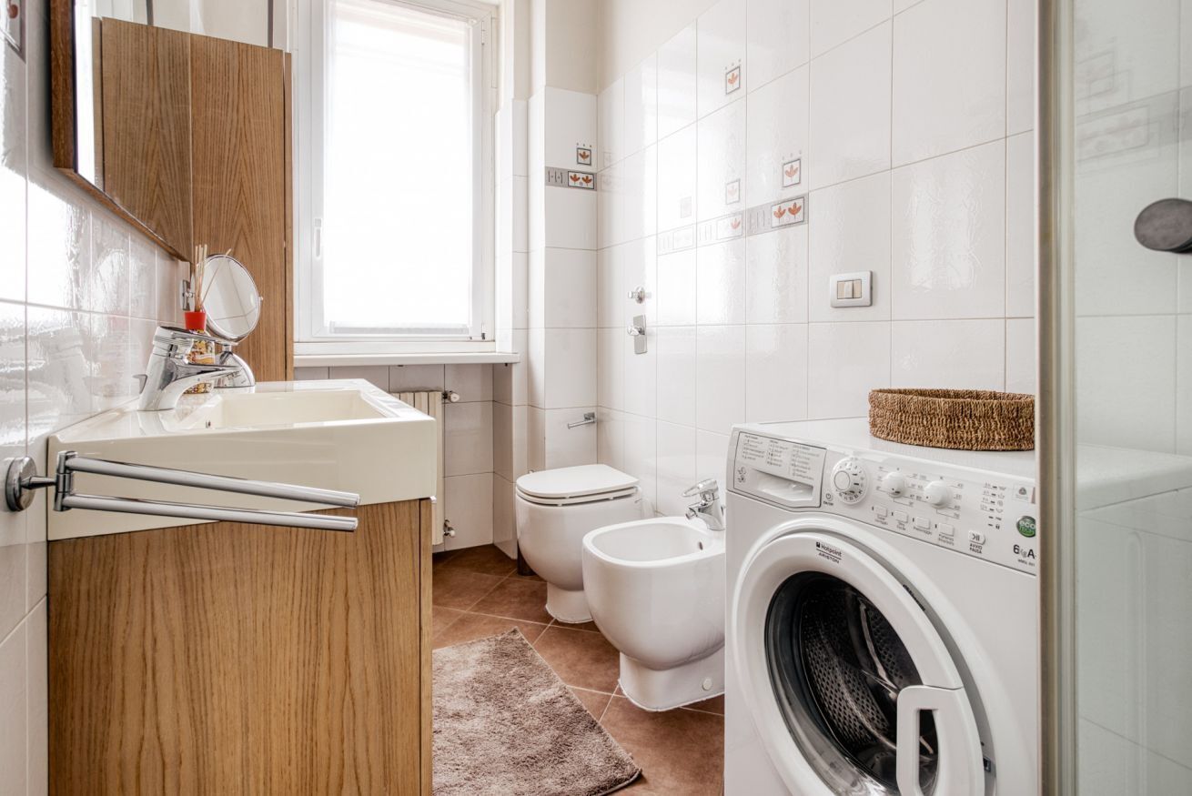 A modern bathroom with a wooden vanity, a washing machine, two toilets, and a window.