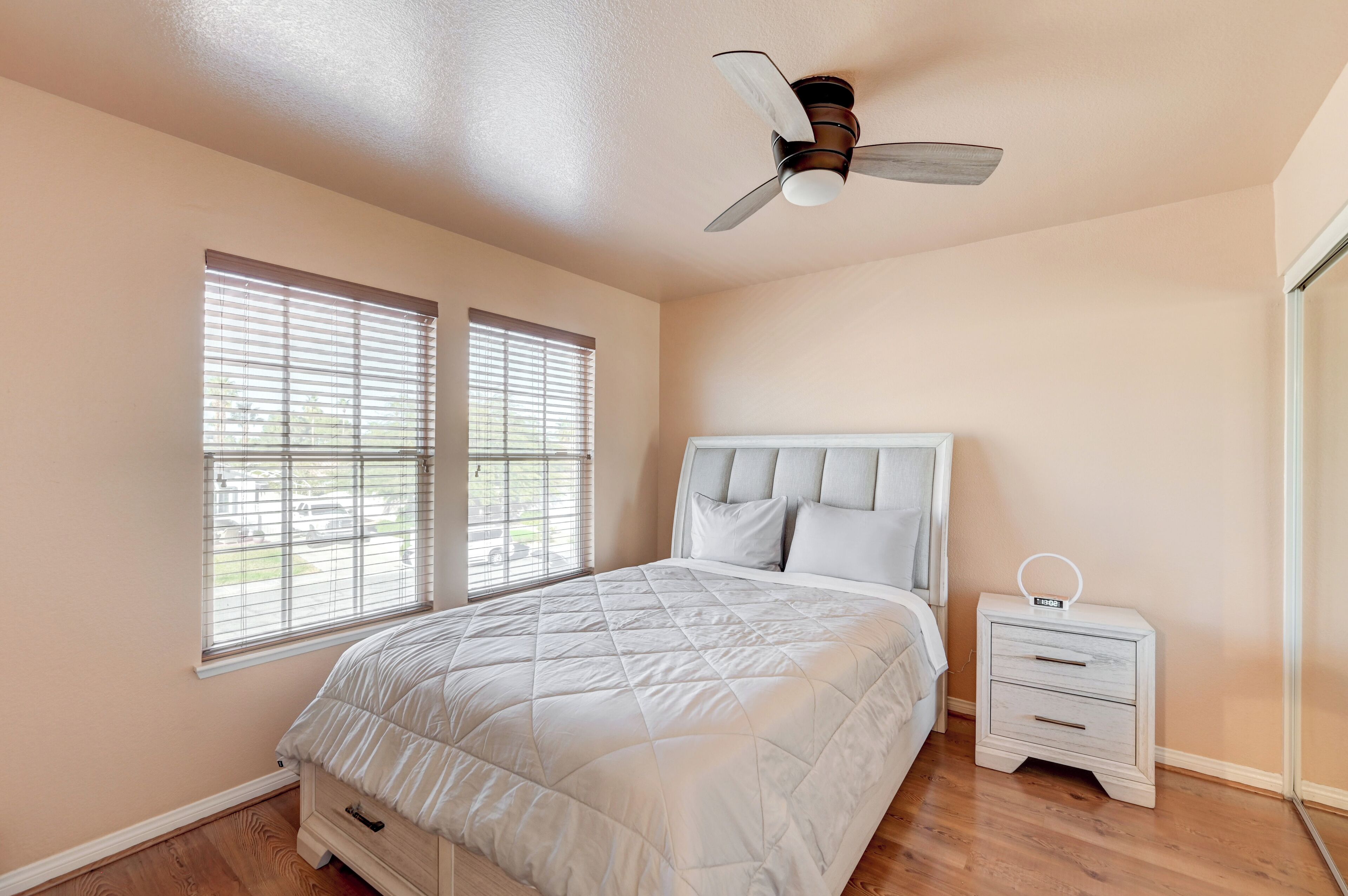 Un dormitorio con cama, dos ventanas con persianas, ventilador de techo, mesita de noche con reloj y mesita auxiliar.