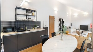 A modern kitchen with dark cabinetry, a white countertop, and a built-in microwave. There is a dining area with a round table and chairs, and a living room with a sofa and a TV. - Stylish Hoxton Duplex – Perfect for Couples & Small Groups (London)
