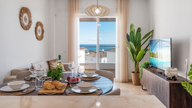 Ett modernt matområde med utsikt över havet, ett matbord dukat med tallrikar, glas och bröd, och en stor växt i hörnet.