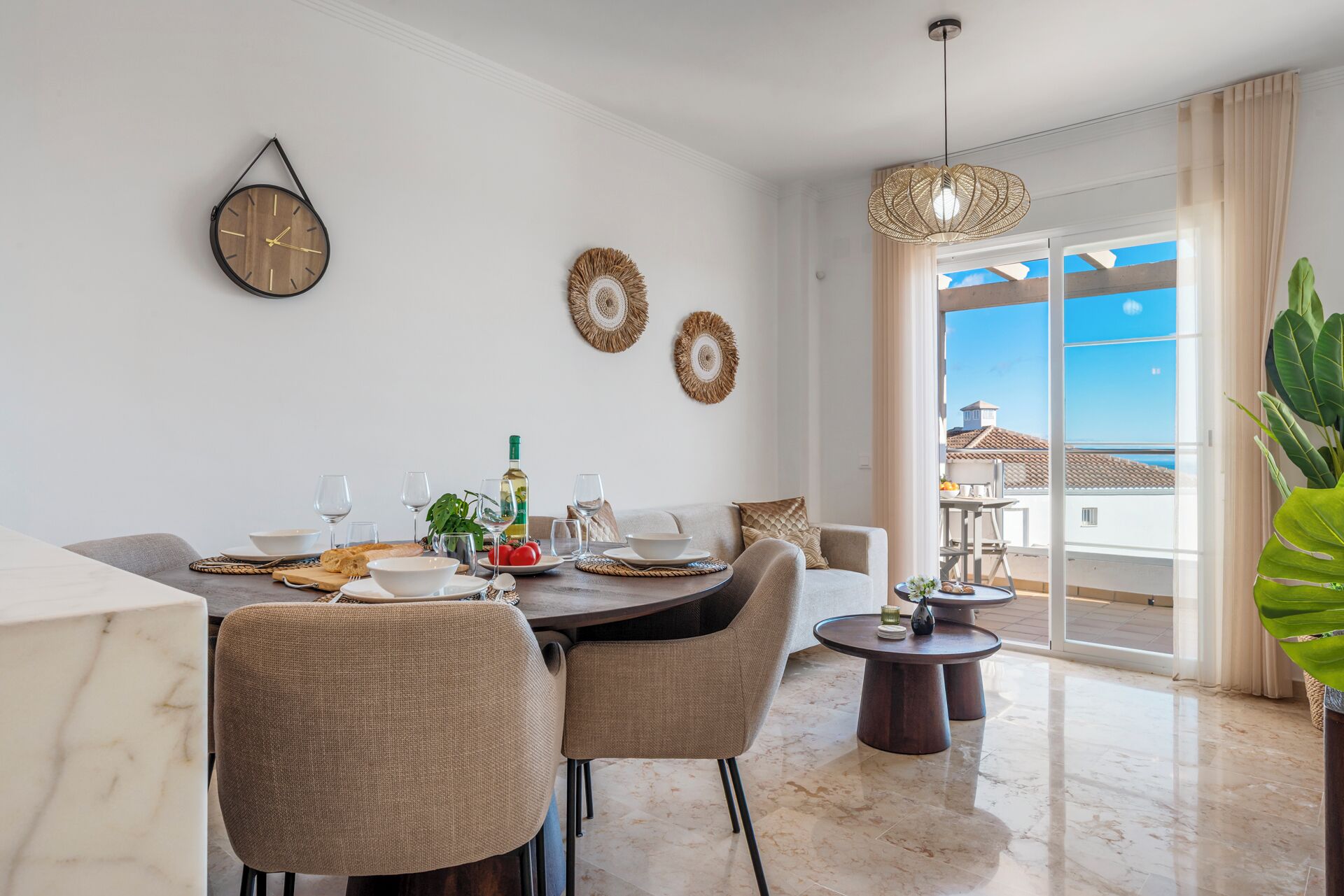Uma área de refeições com uma mesa posta para quatro pessoas, um relógio na parede e vista para o mar através de grandes janelas.