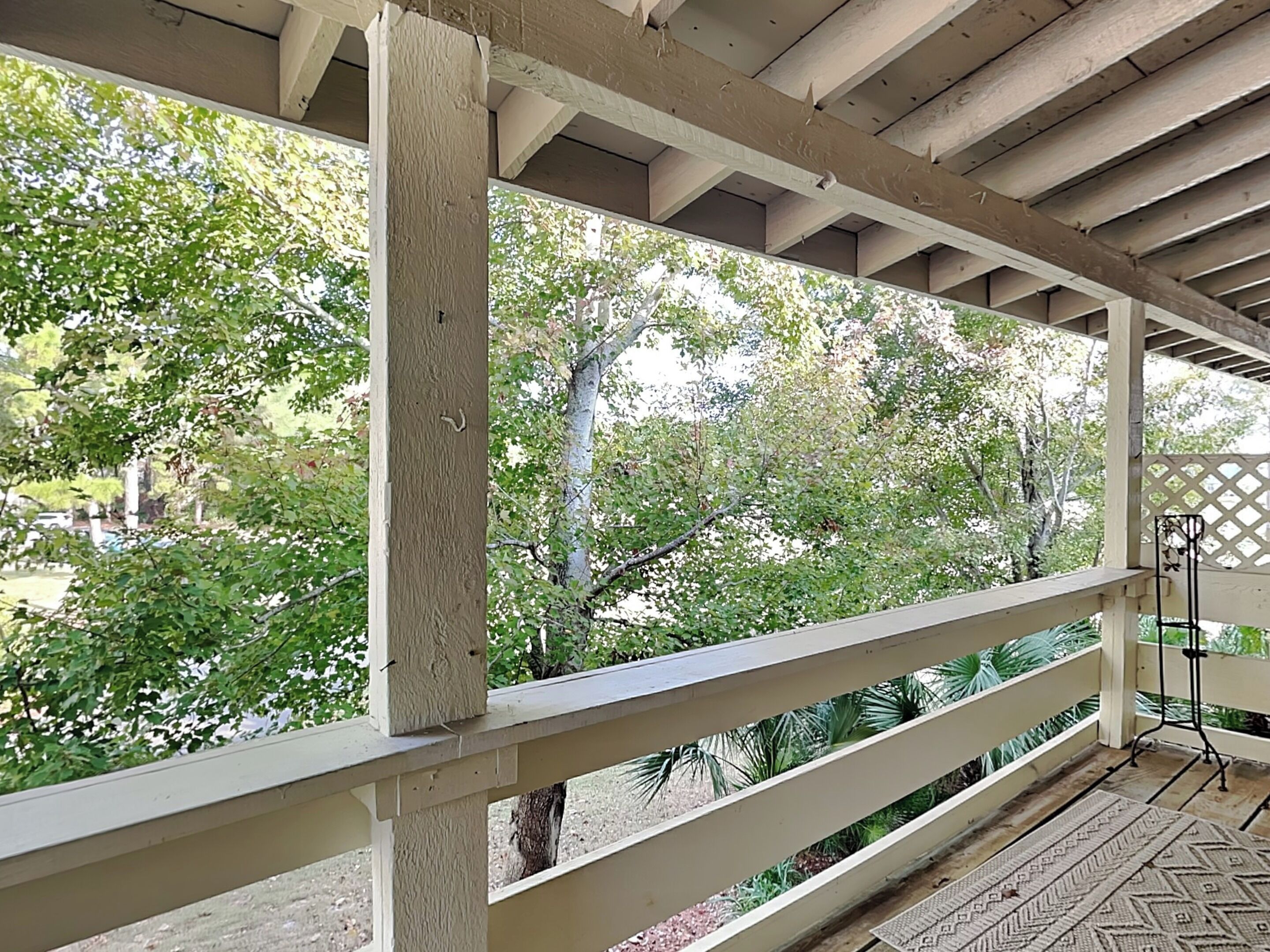A covered outdoor area with a wooden railing, a patterned rug, and a view of trees and greenery.
