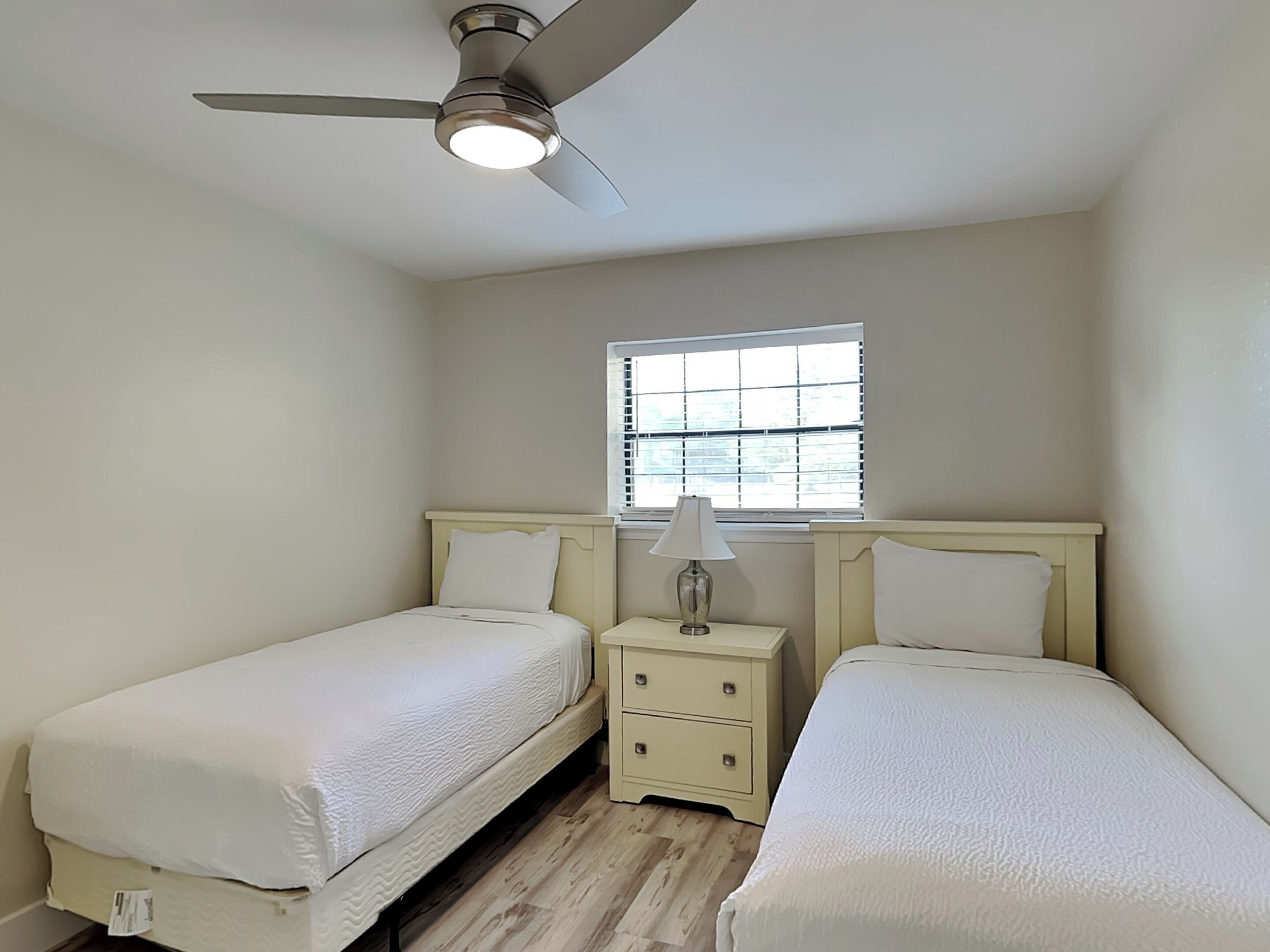 A bedroom with two single beds, a nightstand with a lamp, a ceiling fan, and a window with blinds.