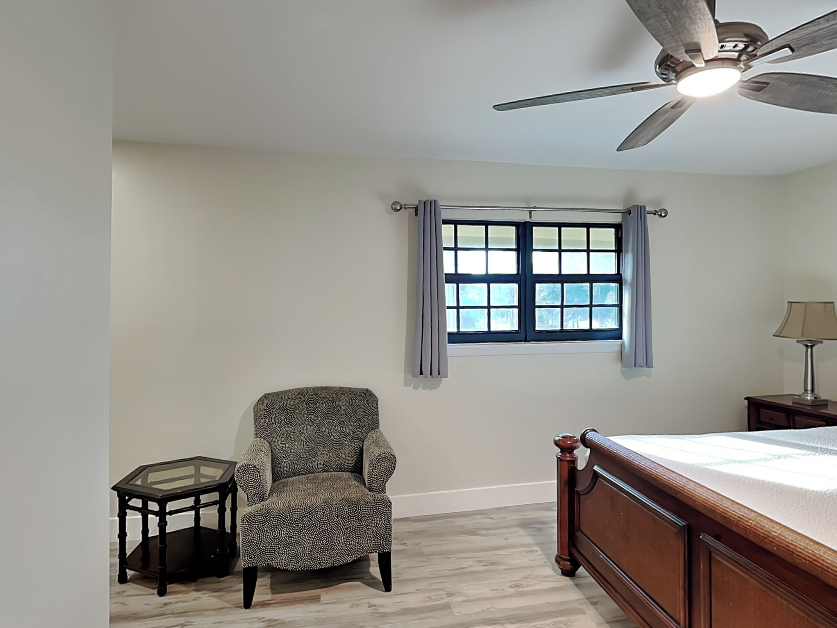 Une chambre à coucher avec un lit, une chaise, une petite table, une fenêtre avec des rideaux et un ventilateur de plafond.